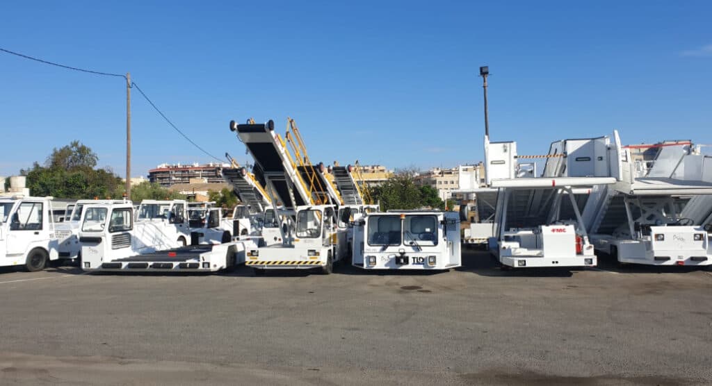 Line of white airport ground support vehicles, including belt loaders and passenger boarding stairs, parked on a sunny tarmac.