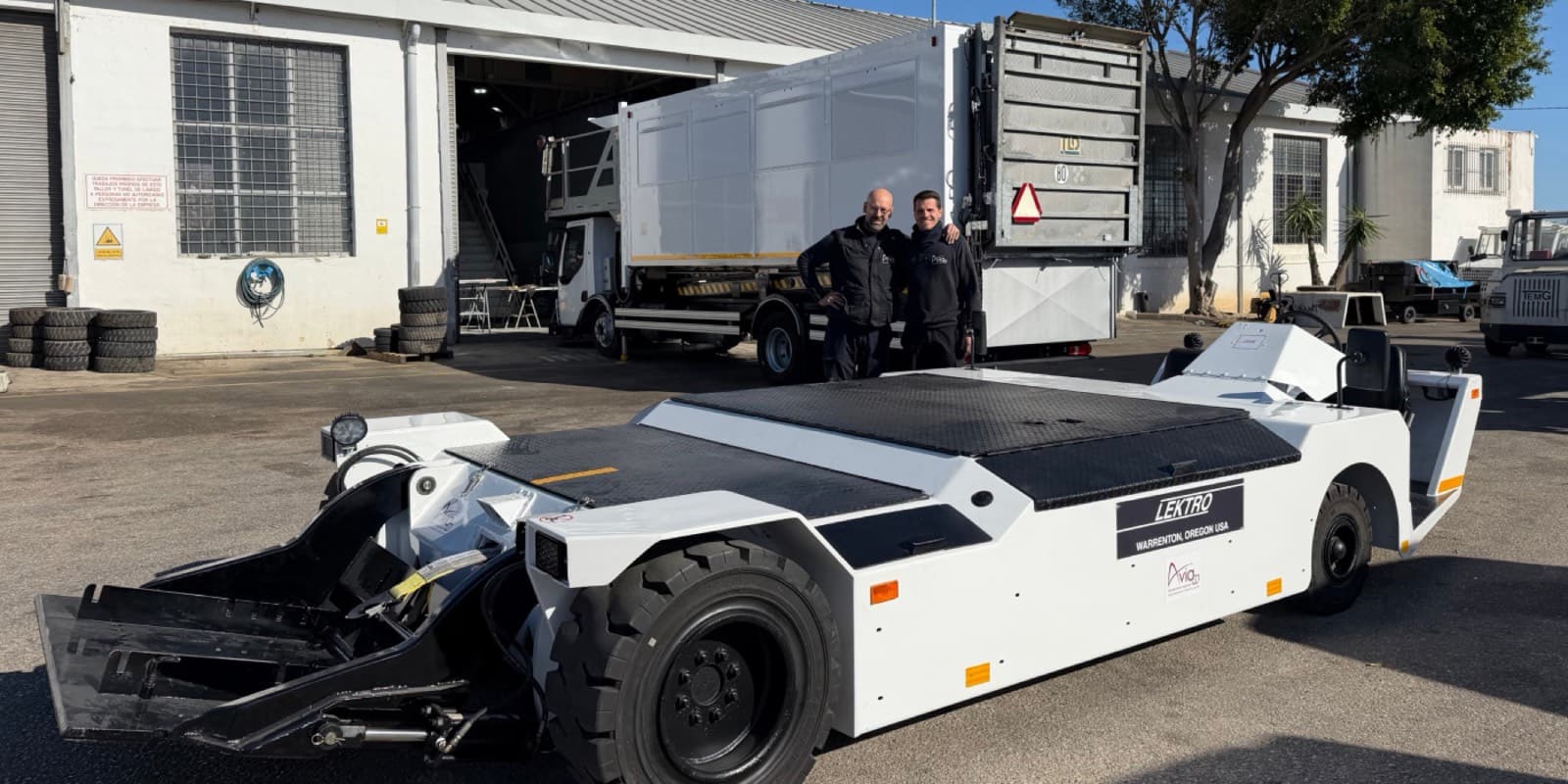 White LEKTRO aircraft tow tractor in a maintenance yard; two technicians stand beside it near a large truck and building.