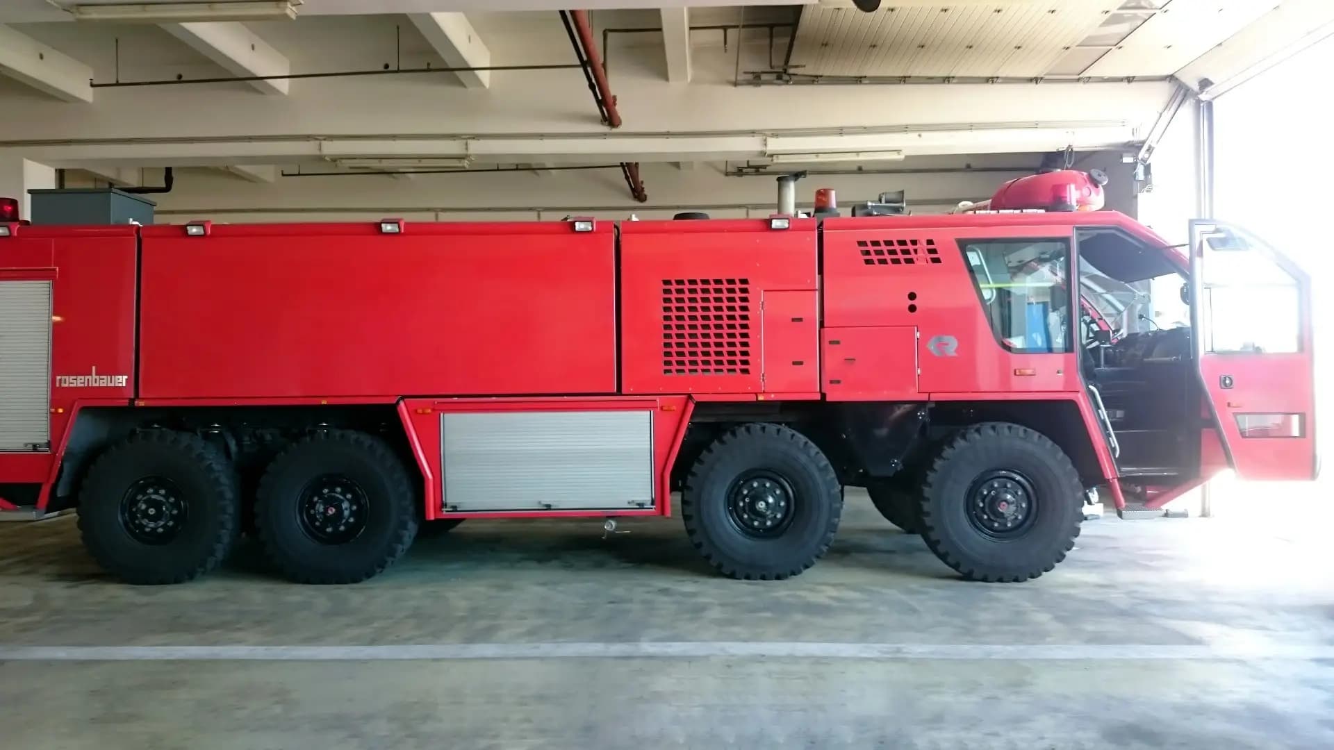 Red fire engine parked inside a garage, view of long side with compartments and large off-road tires; cab door open revealing seating inside.