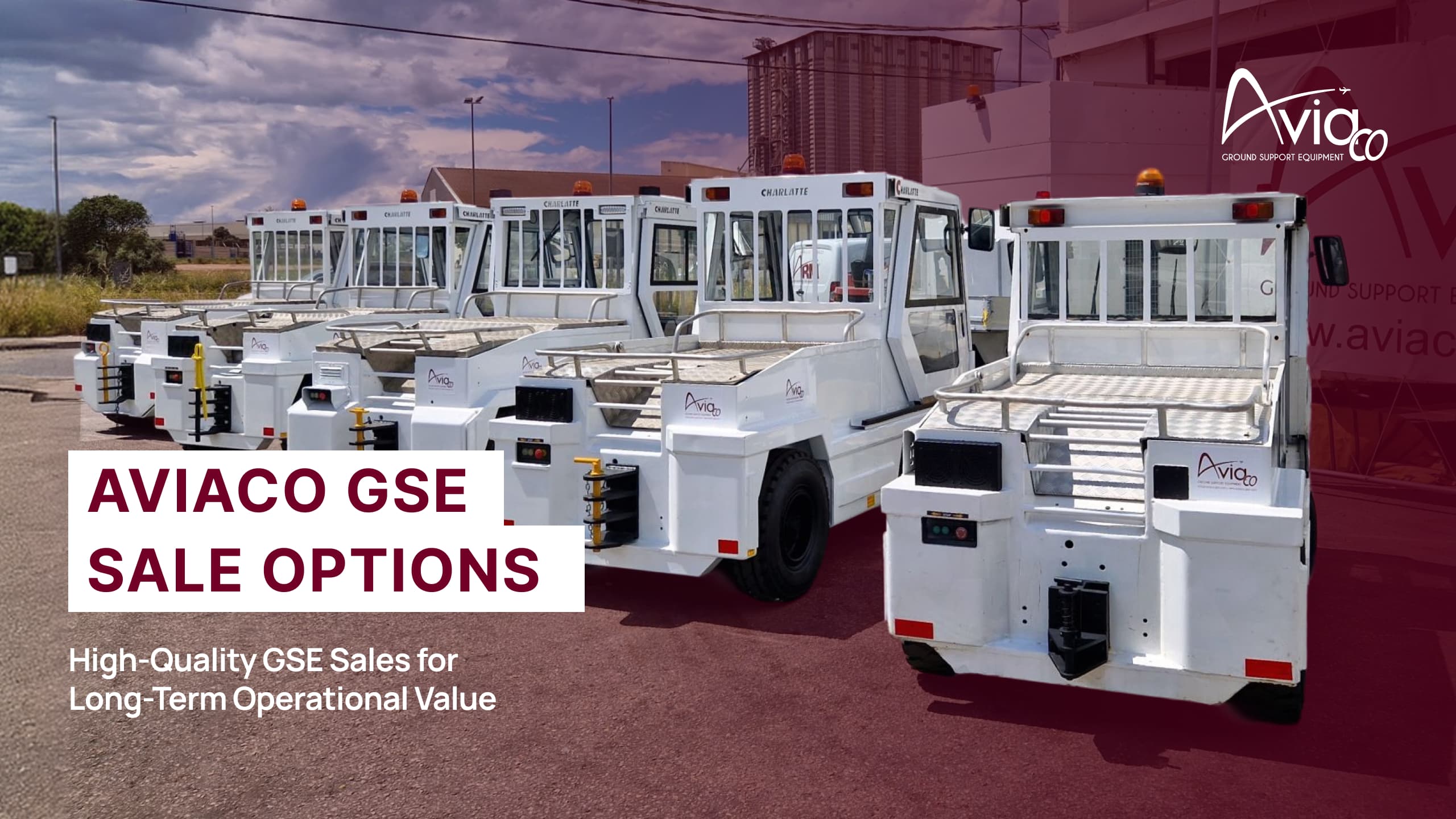 Line of white electric tug vehicles parked outside a warehouse, facing left in a row under a blue sky.