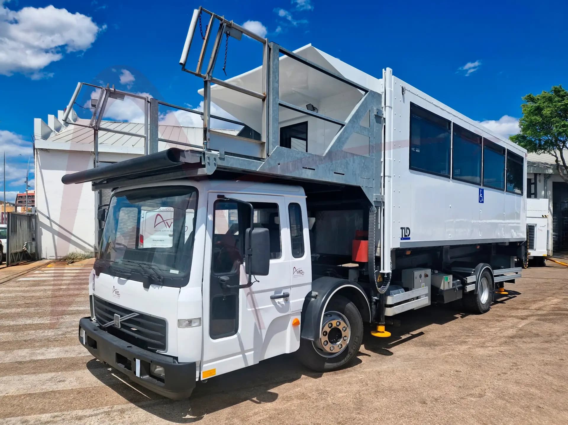 White mobile service unit on a truck with a metal rooftop framework and large side windows, parked on a sunny lot. The wheelchair-accessible sign is visible on the side.