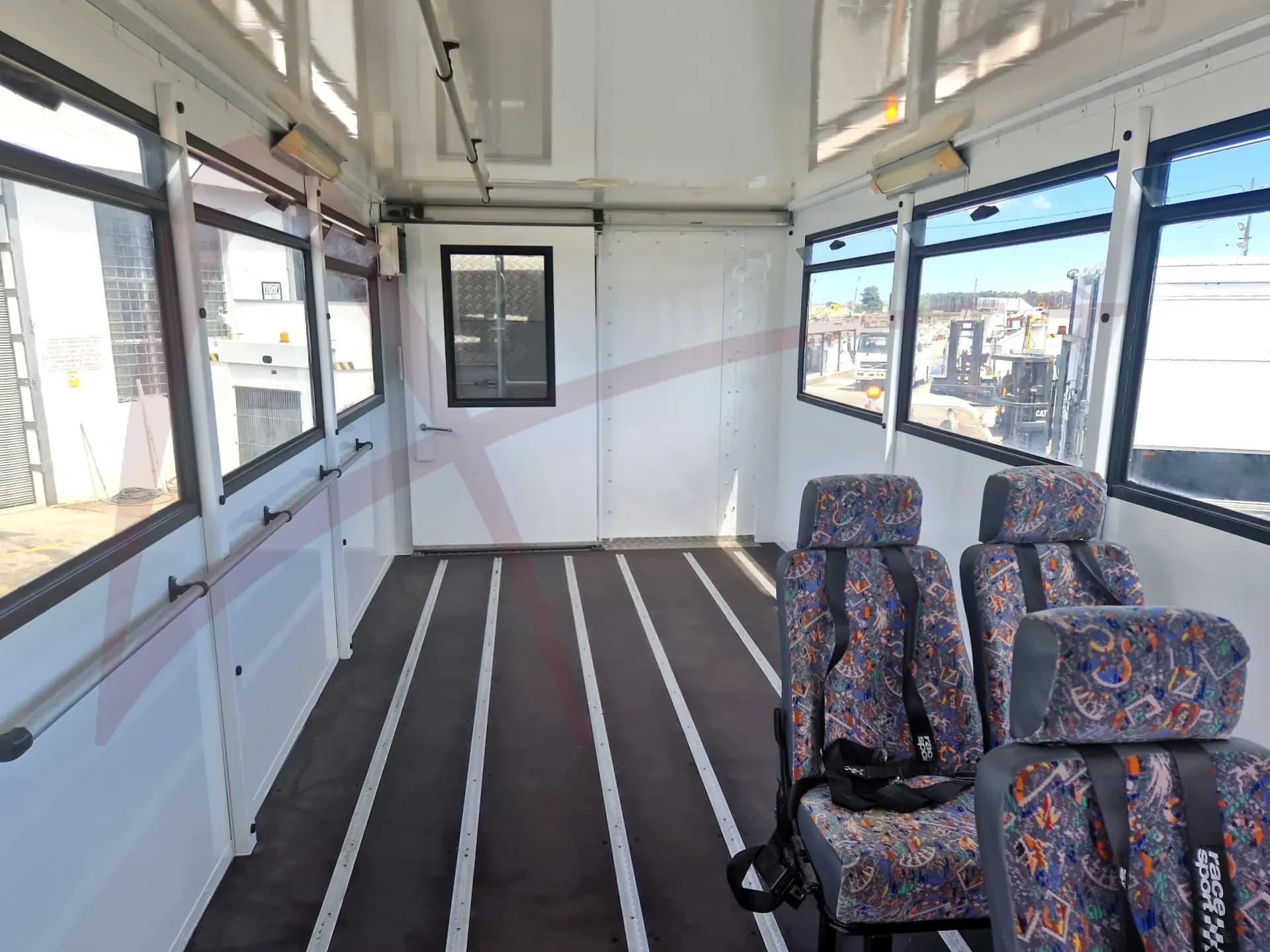 Interior of a white passenger shuttle with patterned fabric seats on the right, large side windows, and a back door visible at the far end.
