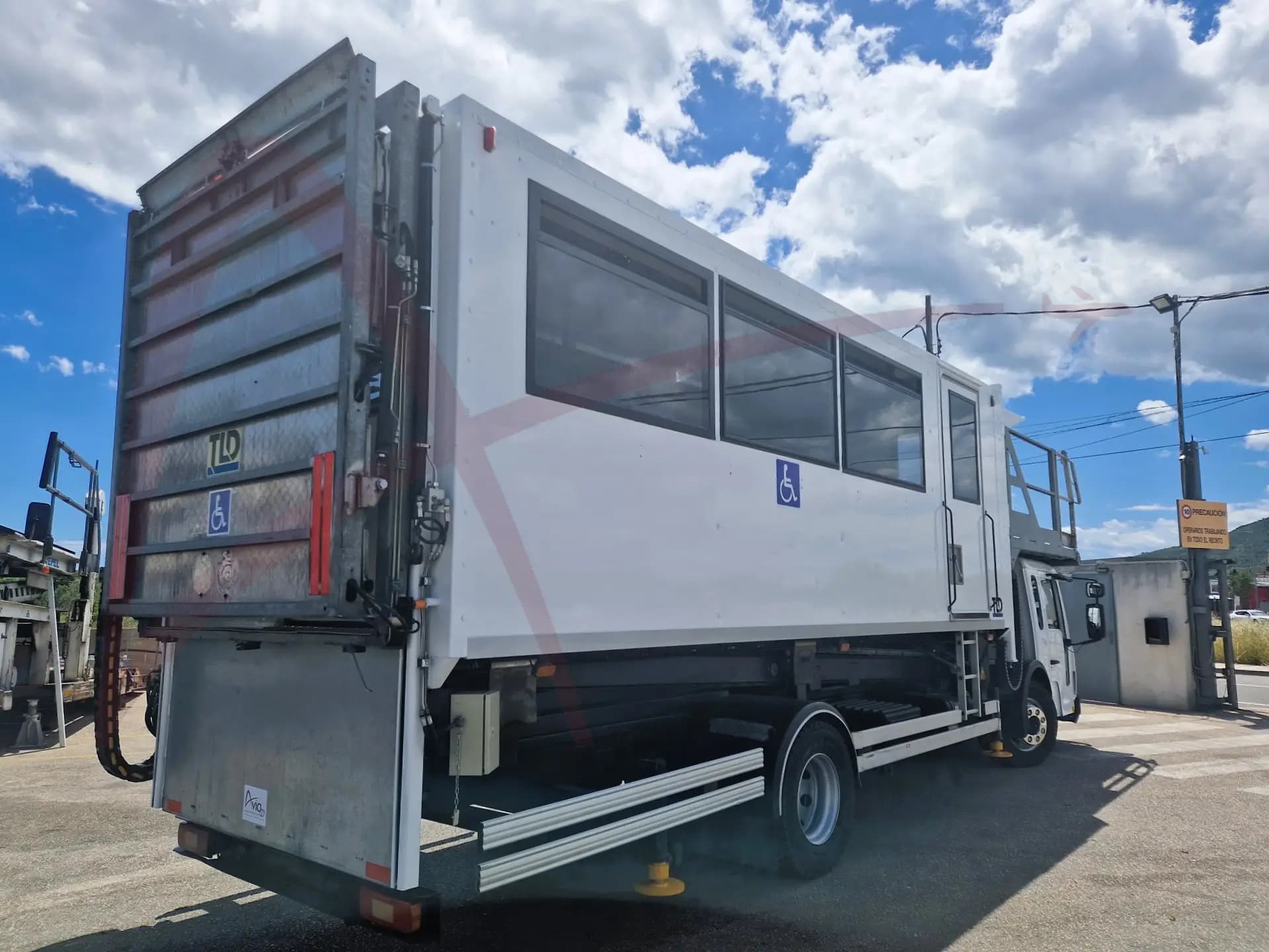 Rear view of a white truck trailer with large side windows and a blue wheelchair accessibility symbol on the side and back doors.