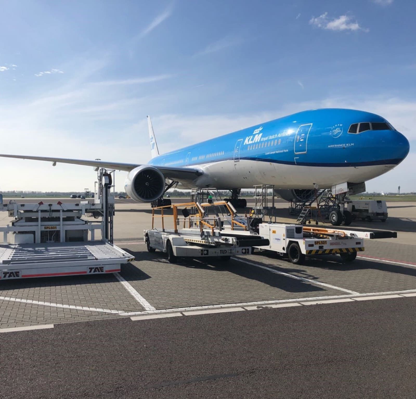 Blue KLM airliner at the gate with ground handling equipment nearby and clear sky above.