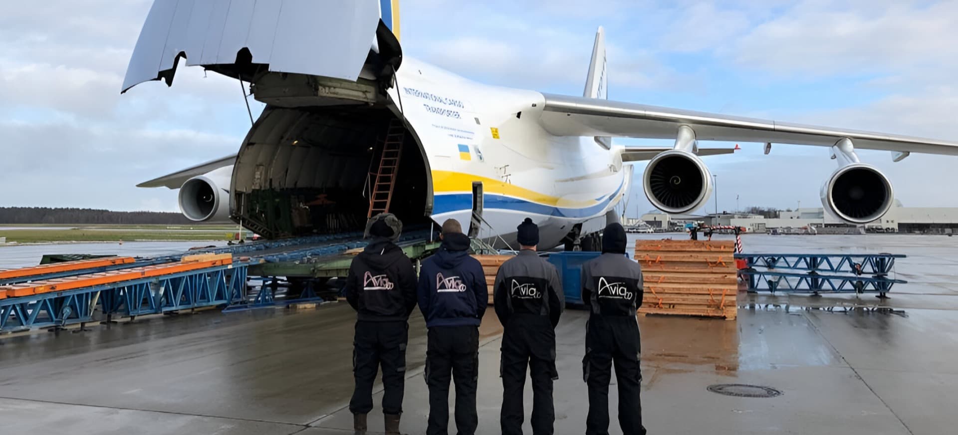 Four workers in dark jackets stand in a line facing a large cargo jet with its rear cargo door open on a wet tarmac, as unloading equipment sits nearby on a sunny day.