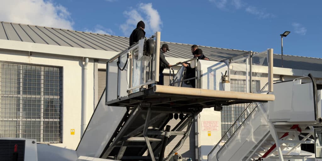 Four workers in dark clothing stand on a raised maintenance platform attached to an industrial structure outside a white building with barred windows.