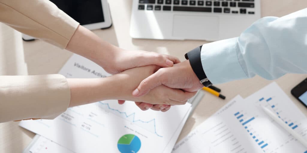Two people shake hands over a desk with business documents and a laptop, symbolizing a partnership or agreement