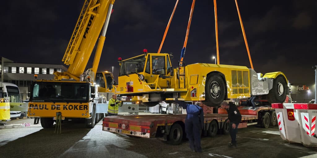Night scene of a large yellow crane lifting a heavy yellow machine onto a flatbed trailer while workers in high‑visibility vests guide the load at an industrial yard.