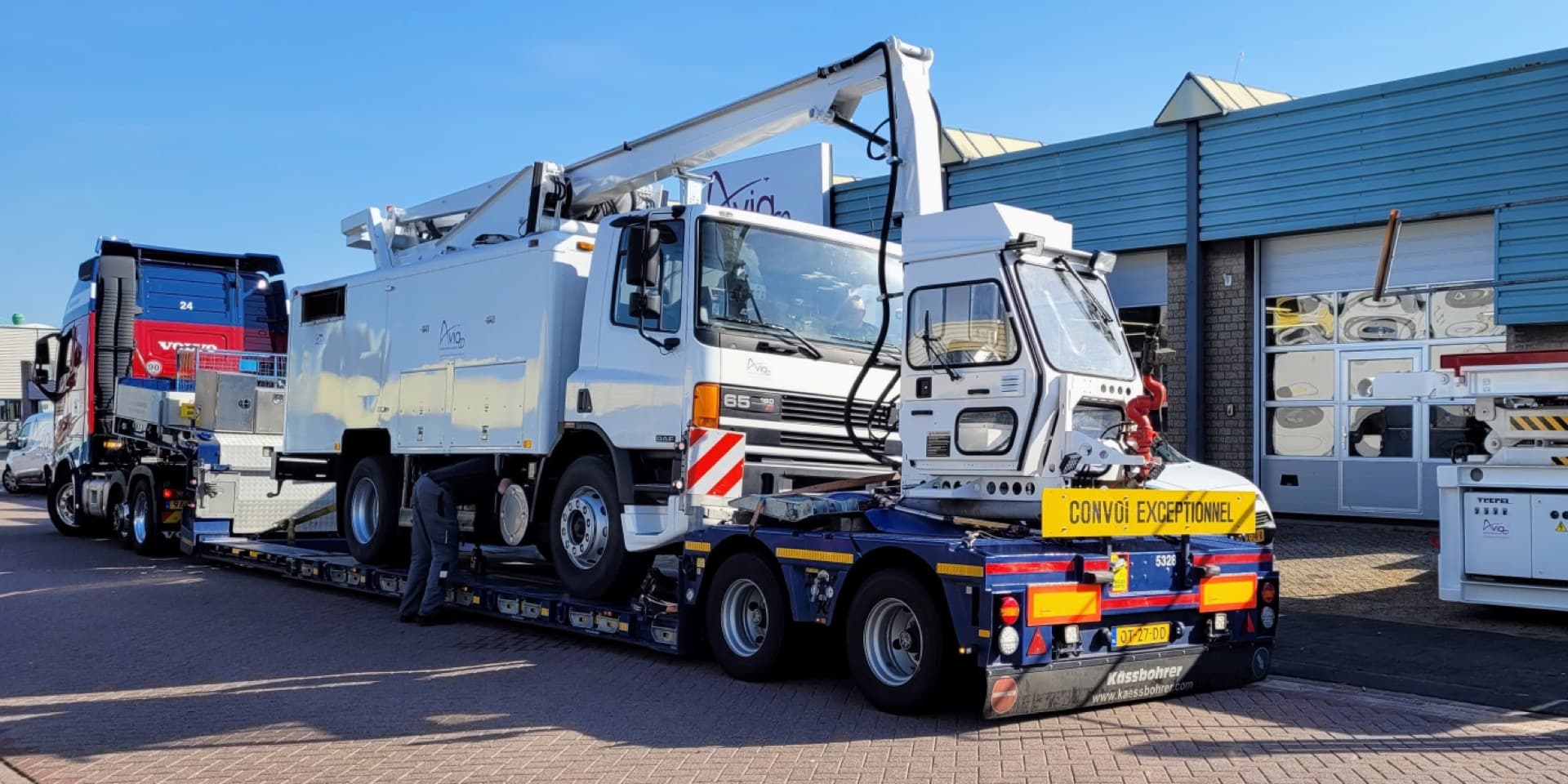 Oversized load truck carrying a large white mobile crane on a lowboy trailer, with a Convoi Exceptionnel sign visible at the rear.