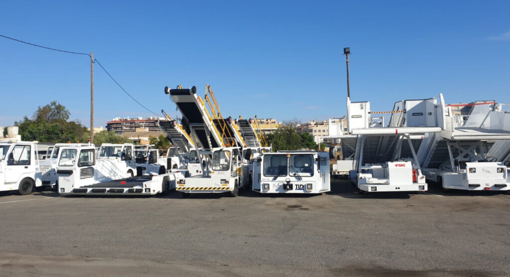 Line of white airport ground support vehicles, including belt loaders and passenger boarding stairs, parked on a sunny tarmac.