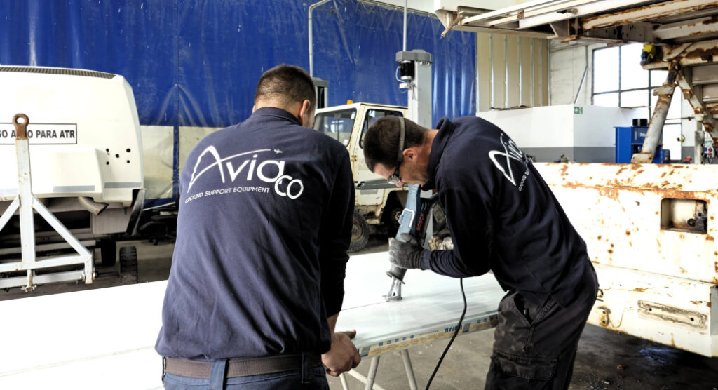 Two workers in matching navy jackets labeled AviaCo cut a large white panel on a worktable inside a maintenance workshop, using a power tool as they lean over the surface.