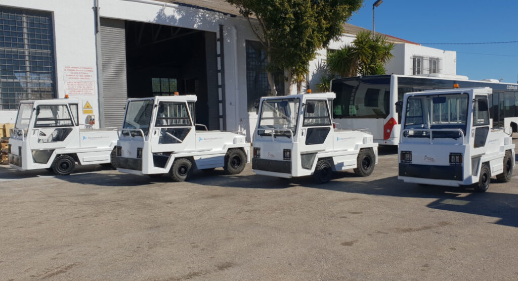 Line of white electric tug vehicles parked outside a warehouse, facing left in a row under a blue sky.