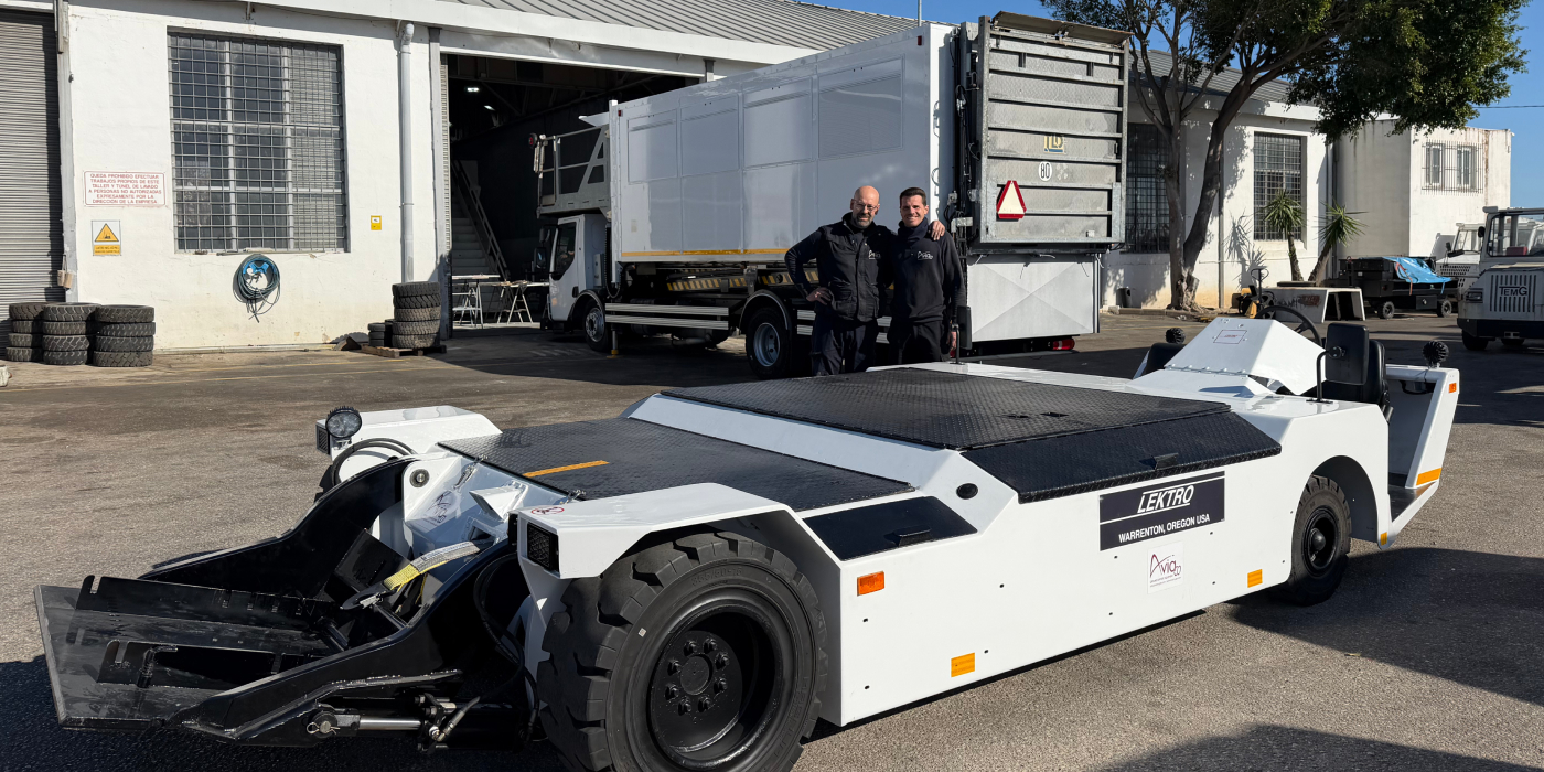 White LEKTRO aircraft tow tractor in a maintenance yard; two technicians stand beside it near a large truck and building.