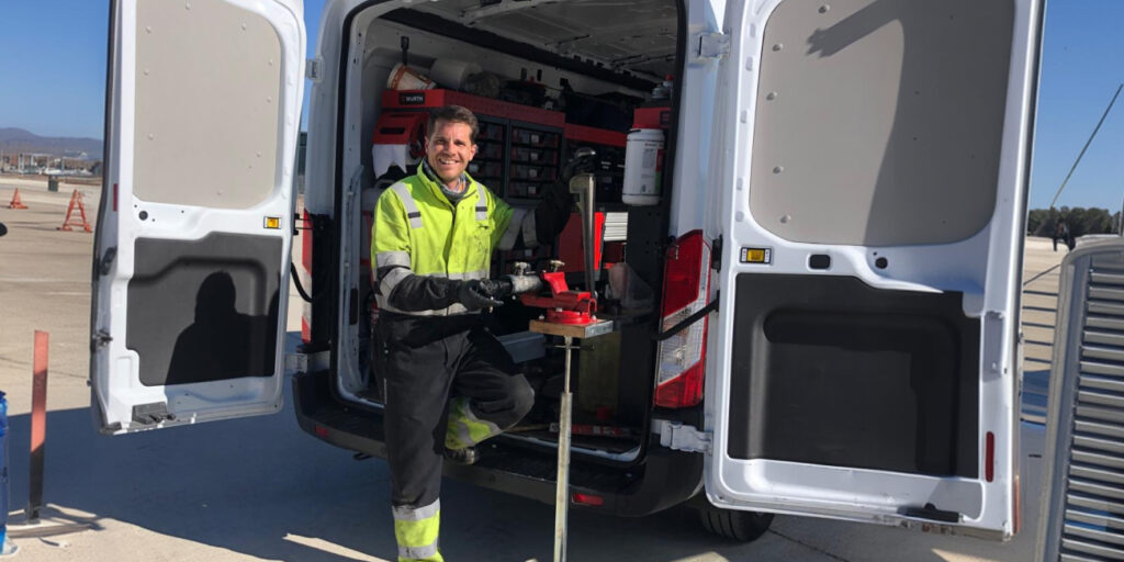 Worker in high‑vis jacket using a tool mounted in the back of a white van, on a tarmac with cones nearby.
