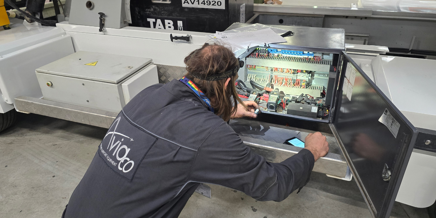 Technician in dark coveralls inspecting wiring inside an open electrical control cabinet on a large industrial machine.