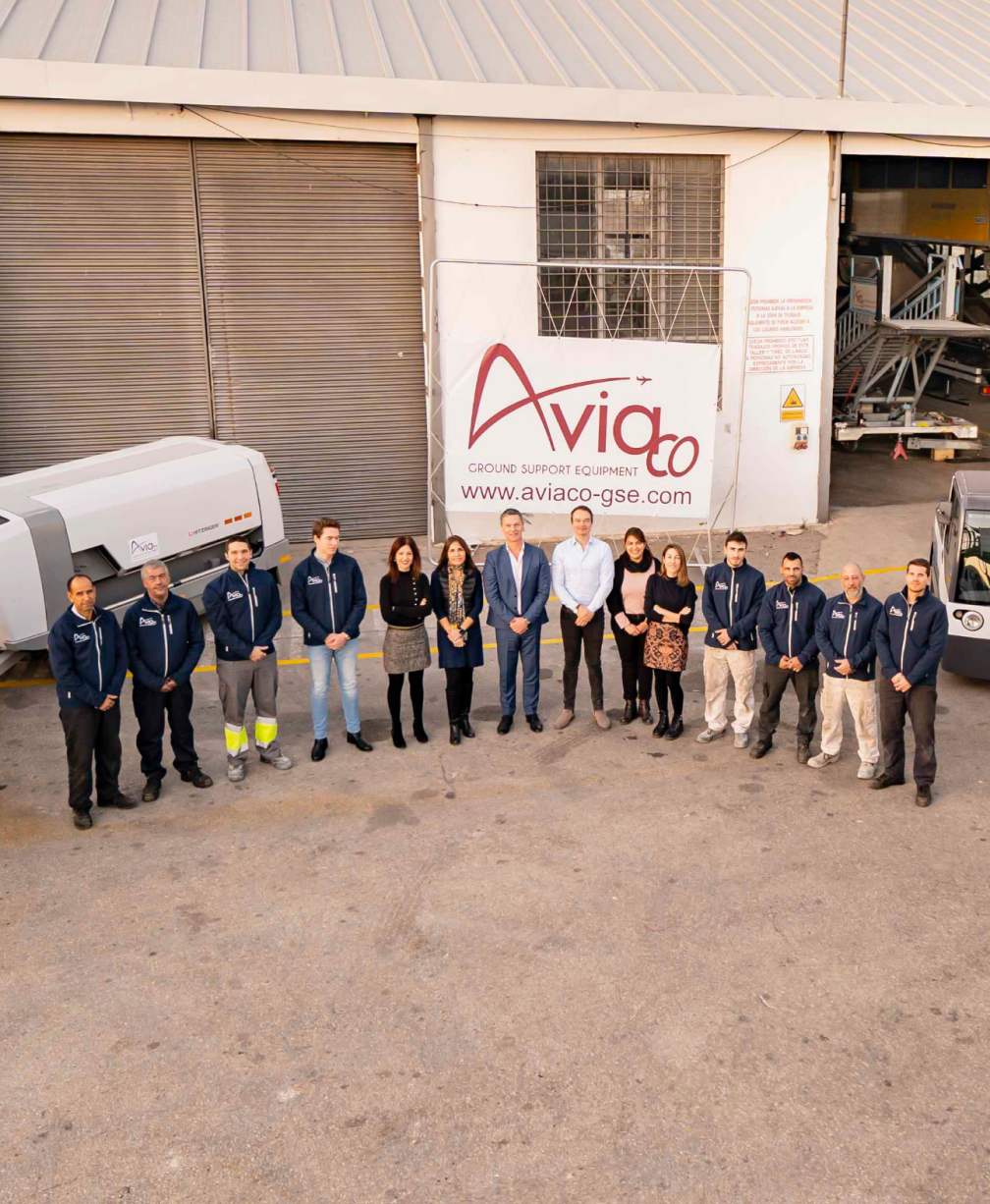 Group of employees standing in a row outside an aviation ground-support equipment facility, in front of a large Aviaco sign with the website listed below it.